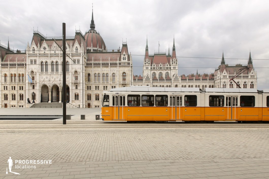 A striking juxtaposition of historic grandeur and modern urban transit, this scene features a grand neo-Gothic parliamentary building with an ornate facade, intricate spires, and a towering central dome. Its white stone exterior, adorned with arched windows and sculptural details, contrasts sharply with the bright yellow tram moving through the foreground. The tram, a classic symbol of urban transportation, runs parallel to the stately structure, emphasizing the blend of historic and contemporary elements. The foreground is dominated by a vast, cleanly paved plaza, adding a sense of openness and scale to the composition. Overhead power lines subtly intersect the frame, reinforcing the city's structured transit system without overwhelming the architectural focus. This location, with its mix of political symbolism and everyday urban life, is ideal for documentary settings, cityscape cinematography, or architectural photography that highlights the contrast between tradition and modernity.