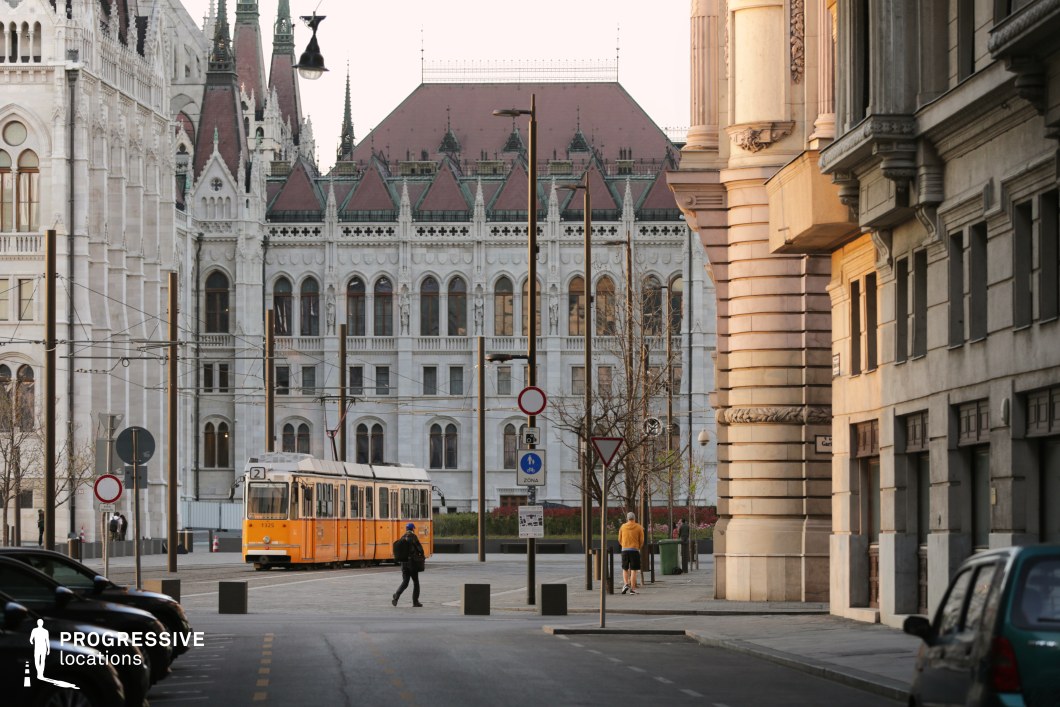 A visually rich urban scene blending historic and modern elements, featuring a grand neo-Gothic parliamentary building with an ornate white facade, pointed spires, and intricate detailing. The foreground is framed by stately, classical buildings with sculpted columns and decorative moldings, adding depth and elegance to the composition. A bright yellow tram moves along the tracks, its vintage aesthetic providing a striking contrast against the surrounding stone architecture. The street is lined with tall, slender streetlights and tram poles, creating vertical elements that enhance the sense of perspective. The mix of architectural grandeur and functional urban design makes this location ideal for cinematic cityscapes, period dramas, or modern urban storytelling, capturing the timeless elegance of a European capital.