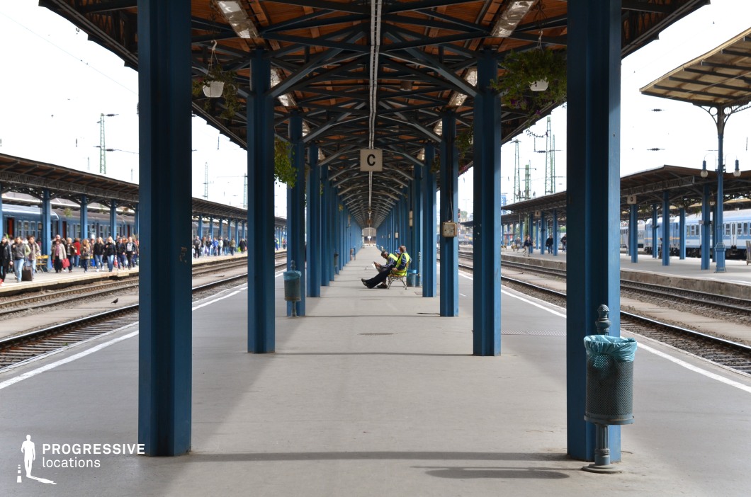 This traditional open-air railway station features a striking linear perspective created by evenly spaced blue steel columns supporting a wooden roof structure. The covered platform extends into the distance, emphasizing depth and symmetry, while multiple train tracks on either side reinforce the sense of movement and transit. The industrial elements, such as metal framework and overhead power lines, contrast with the warm wooden tones of the roof, creating a balanced blend of functionality and aesthetics. The surrounding platforms, partially visible in the background, add to the station's structured, organized atmosphere. This location is ideal for capturing the energy and rhythm of travel while offering a timeless setting for transportation-focused visuals.