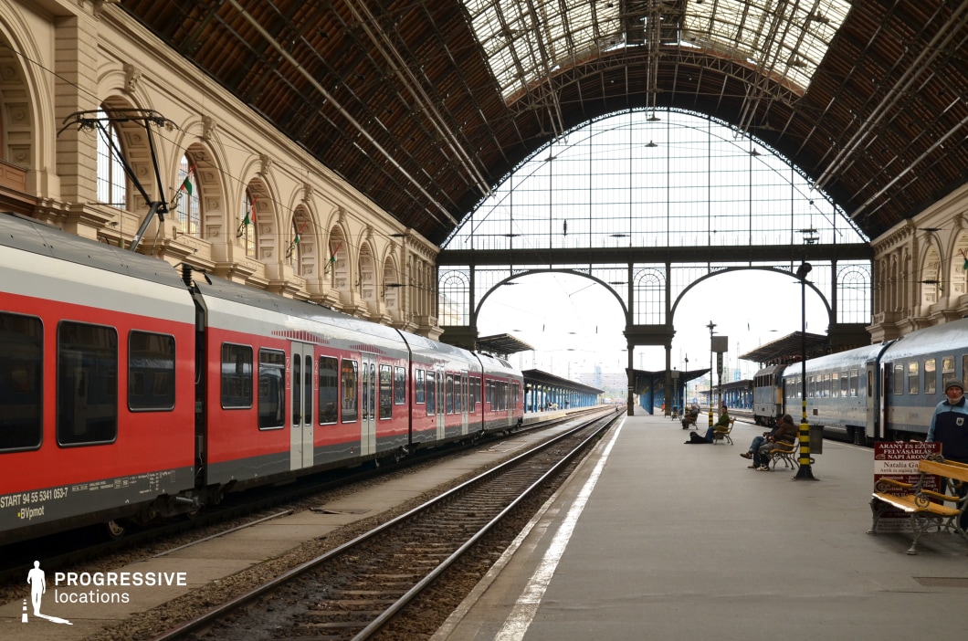 This impressive railway station blends classical architecture with industrial-era design, featuring a vast arched glass roof supported by an intricate metal framework. The platform is flanked by historic facades adorned with elegant arched windows and decorative moldings, while modern trains contrast against the traditional setting. The high ceiling and expansive glazing allow ample natural light to illuminate the platform, enhancing the station's grandeur and openness. The steel beams and symmetrical arches create a strong visual rhythm, drawing attention toward the tracks leading into the distance. This location offers a dynamic mix of old-world sophistication and contemporary functionality, making it ideal for period dramas, travel-related visuals, or cinematic urban transit settings.