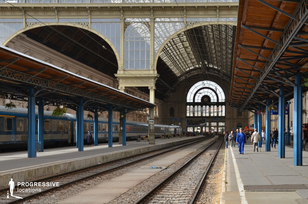 This expansive railway station showcases a striking blend of classical and industrial architecture, featuring a towering arched glass roof supported by intricate metalwork. The symmetrical design draws the eye towards the grand central facade, where large arched windows allow natural light to flood the station, highlighting the elegant masonry and refined details. The platform is framed by steel and wood canopies, adding a layer of depth and structure, while the railway tracks lead into the distance, emphasizing the sense of movement. The combination of historic elegance and functional modernity makes this location ideal for capturing the timeless charm of grand railway travel and urban transit hubs.