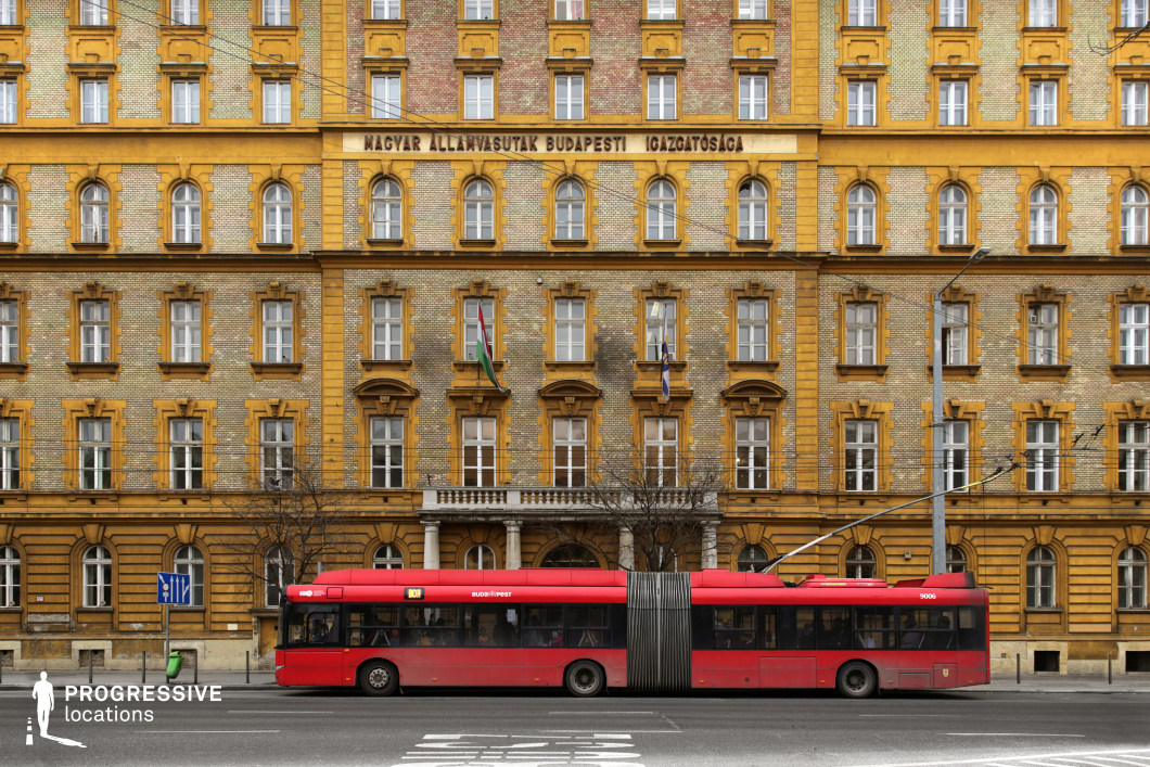 This striking neoclassical-style building features a richly detailed yellow brick facade, adorned with symmetrically arranged arched windows and decorative moldings. The architectural design emphasizes elegance and grandeur, with contrasting brickwork and intricate stone elements that highlight its historic character. The facade is punctuated by a central balcony, adding depth to the structure. In front of the building, a red articulated trolleybus moves along the street, introducing a dynamic element that contrasts with the rigid symmetry of the architecture. The overhead power lines and urban infrastructure enhance the metropolitan atmosphere, making this location ideal for capturing the intersection of historical architecture and modern city life.