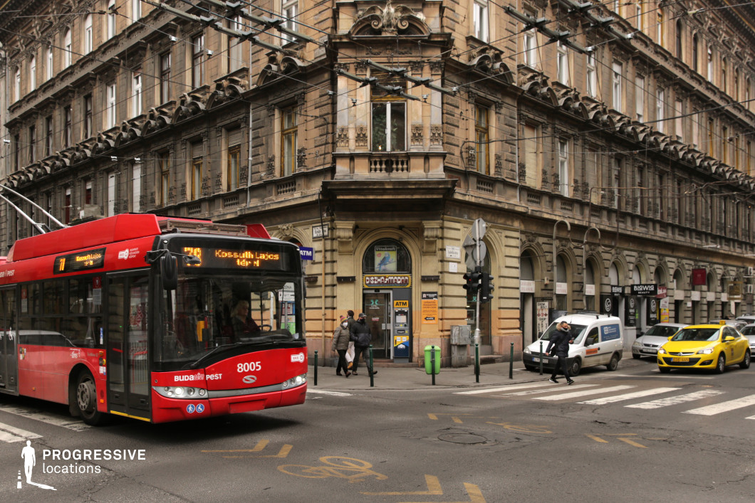 This vibrant city intersection is framed by a grand historic corner building, featuring intricate stonework, arched entryways, and decorative balconies that reflect classic European architectural influences. The facade’s aged patina adds to its timeless character, while the dense network of overhead trolley wires enhances the urban atmosphere. A red trolleybus navigating the curve of the street provides a striking contrast to the muted tones of the architecture, reinforcing the dynamic interaction between historic structures and modern transit. The layered streetscape, featuring a mix of vehicles and pedestrian activity, contributes to the scene’s authentic metropolitan energy, making it an ideal setting for urban storytelling and cityscape compositions.