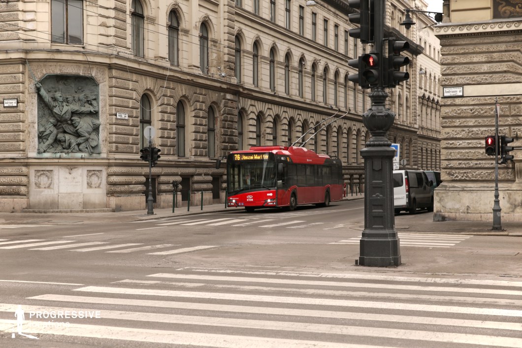 This stately city intersection is framed by grand limestone-clad buildings with ornate architectural details, featuring arched windows, intricate cornices, and a sculptural relief embedded in the facade. The textured stonework and classical design elements lend the location a distinguished, old-world charm. A red trolleybus moves along the street, contrasting against the neutral tones of the historic buildings while highlighting the fusion of past and present in the cityscape. The structured composition, including the bold zebra crossing in the foreground and decorative street fixtures, enhances the sense of order and symmetry. This setting is ideal for capturing the elegance of historic urban environments with a strong metropolitan character.