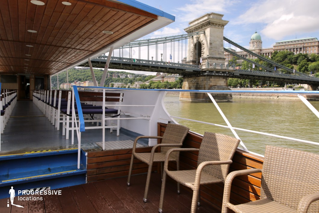 This open-air riverboat deck provides a panoramic view of a historic suspension bridge and grand architectural landmarks along the waterway. The wooden flooring and ceiling, combined with a mix of seating areas, create a relaxed yet structured environment ideal for sightseeing. The bridge’s massive stone pillars and intricate steel framework form a dramatic focal point, while the background cityscape, featuring classical buildings atop a green hillside, adds depth and elegance to the scene. The river’s reflective surface enhances the visual balance between natural and built elements, making this location ideal for capturing the charm of riverfront travel and iconic cityscapes.