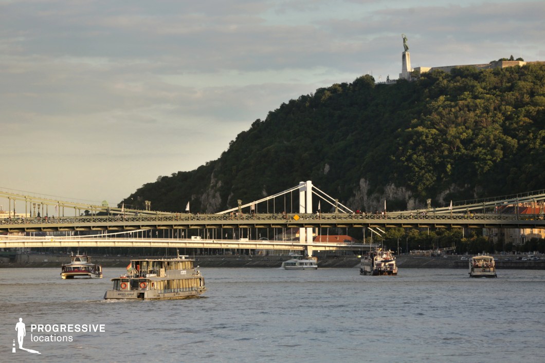 This picturesque riverside scene features multiple bridges spanning a wide, gently flowing waterway, with boats navigating the current, adding a dynamic element to the composition. The bridges, showcasing a mix of industrial and modern design, contrast against the lush, forested hillside rising in the background. Atop the hill, a prominent statue stands as a recognizable landmark, reinforcing the area's historical and cultural significance. The layering of natural and built elements, combined with the movement of the boats, creates a balanced and visually engaging composition. This setting is ideal for capturing the harmony between urban infrastructure and the surrounding natural landscape.