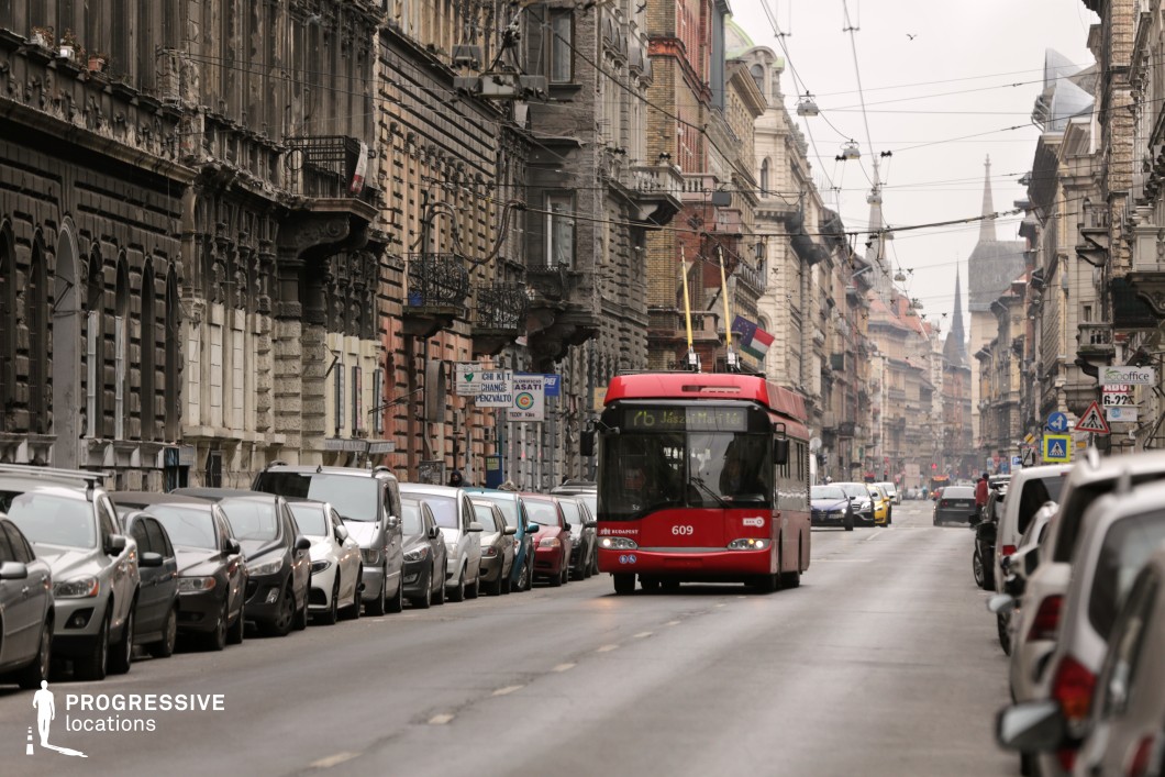 This atmospheric city street is lined with historic buildings featuring ornate facades, wrought-iron balconies, and intricate architectural details that evoke a European old-world charm. The mix of preserved and weathered exteriors enhances the textured, layered aesthetic of the setting. A red trolleybus moves through the scene, framed by the dense network of overhead wires that emphasize the urban transit infrastructure. The perspective of the street extends into the distance, leading towards church spires and additional architectural landmarks, adding depth and a sense of history. This location is ideal for capturing the authentic character of a historic city with a blend of tradition and modern movement.