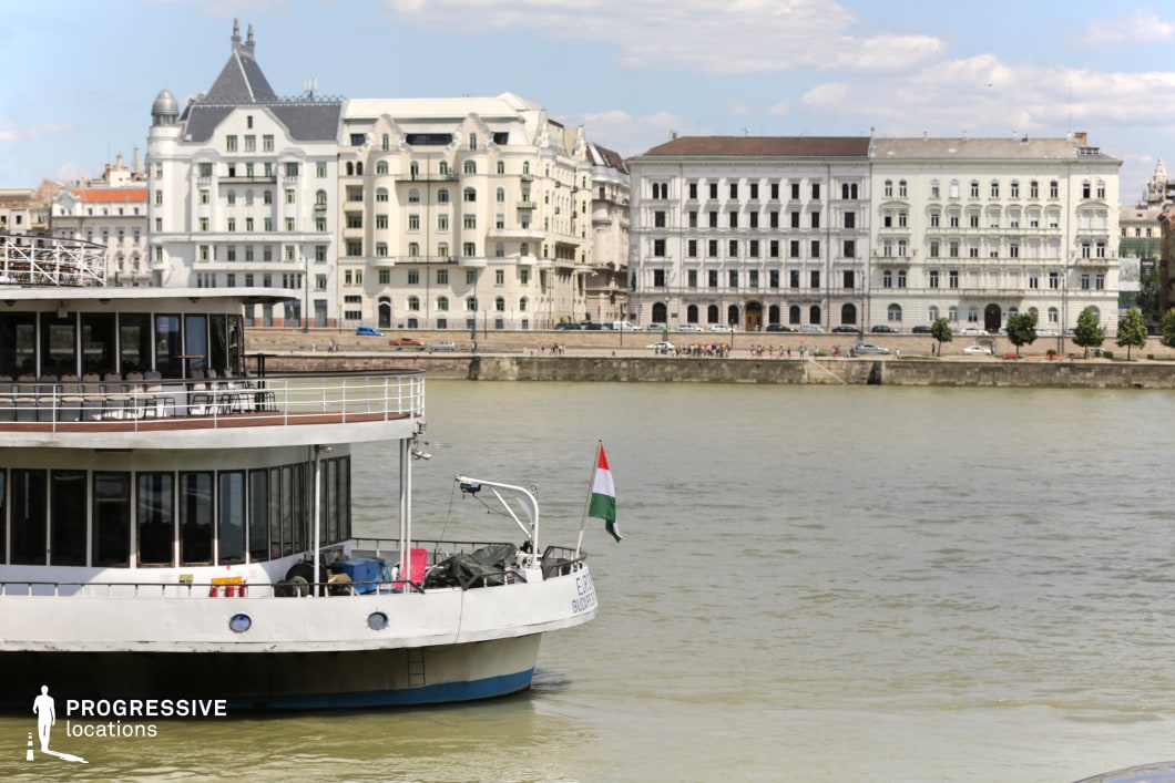 This scenic riverfront captures the harmony between historic architecture and modern waterway transit. A large, multi-deck cruise ship with an open-air seating area is positioned in the foreground, adding depth to the composition. The background showcases a row of grand, neoclassical and Art Nouveau-style buildings with intricate facades, arched windows, and decorative elements, emphasizing the city’s rich architectural heritage. The river acts as a natural divider, reflecting the surrounding structures and reinforcing the sense of openness. The combination of water, elegant urban design, and the presence of the ship makes this location ideal for capturing the sophisticated ambiance of riverside city life.