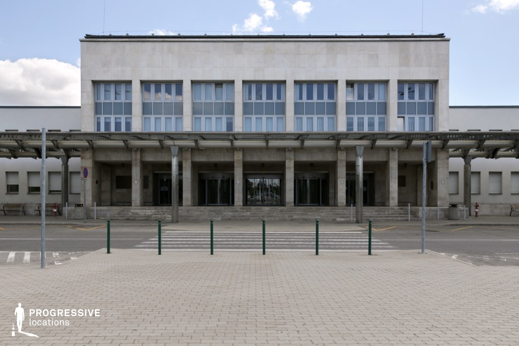 This imposing airport entrance reflects a functionalist architectural style, characterized by its rigid symmetry, clean lines, and minimalist facade. The building is clad in light-colored stone, with large rectangular windows that emphasize its utilitarian aesthetic. A covered walkway with steel supports extends across the entrance, adding depth and structure while reinforcing the practical design. The paved foreground, featuring a pedestrian crossing and evenly spaced bollards, enhances the sense of order and control. This location is ideal for capturing the stark efficiency of mid-century modern infrastructure, making it suitable for settings that require a formal, institutional backdrop.