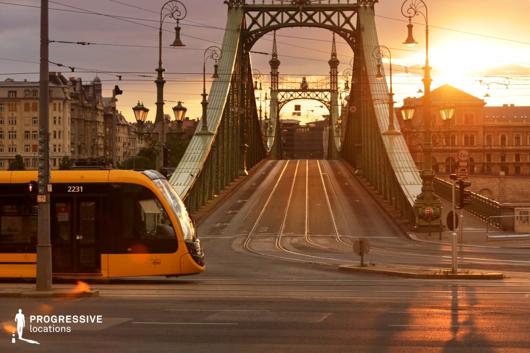 This striking urban scene captures the contrast between historic and contemporary elements, featuring an ornate steel bridge with intricate detailing and decorative lampposts stretching across the cityscape. The tram lines seamlessly integrate into the bridge’s structure, emphasizing the fusion of past and present in urban transport. A sleek, modern yellow tram moves through the foreground, adding a dynamic element that contrasts with the bridge’s classical design and surrounding historic buildings. The structured symmetry of the bridge, combined with its elegant curves, creates a visually engaging composition that highlights the city’s rich architectural heritage and efficient public transit system.