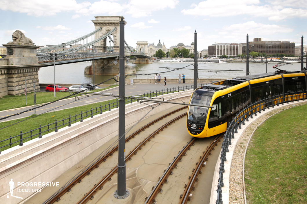 This dynamic cityscape blends historic and contemporary elements, featuring a modern yellow tram curving along a sleek track beside a grand suspension bridge. The stone bridge, adorned with lion sculptures and intricate ironwork, serves as a striking focal point, emphasizing the city’s architectural heritage. The riverbank, lined with pathways and green spaces, creates a scenic contrast to the urban infrastructure, while the background showcases a mix of classic and modern buildings. Overhead tram wires add a structured, industrial element, reinforcing the seamless integration of past and present transportation networks. This location is ideal for capturing the elegance of historic landmarks within a vibrant urban setting.