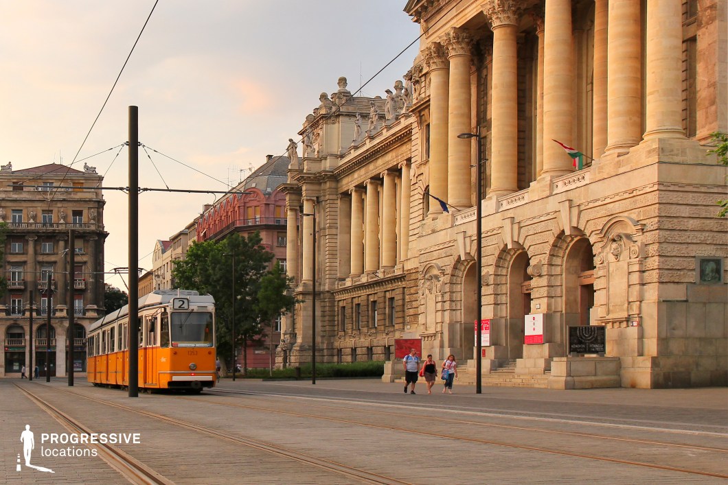 This elegant cityscape captures a grand neoclassical building with towering columns, intricate stonework, and sculptural details along its ornate facade. The imposing structure, bathed in warm light, stands as a testament to classical architectural grandeur. A vintage yellow tram moves along embedded tracks in the foreground, adding a dynamic contrast to the building’s timeless elegance. The paved plaza and surrounding historic buildings create a harmonious blend of old-world sophistication and urban functionality. The scene’s balanced composition, with its interplay of transit and monumental architecture, makes it an ideal location for capturing the charm of a historic European city.
