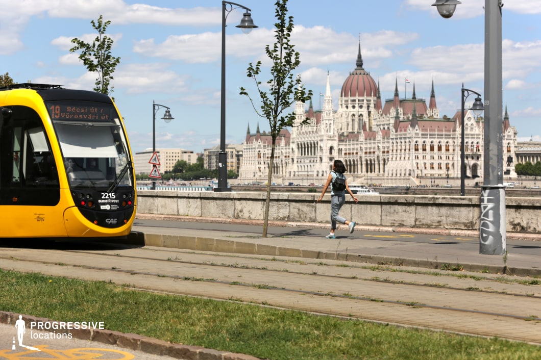 This vibrant cityscape captures the perfect blend of historic grandeur and contemporary urban transit, featuring a sleek yellow tram traveling along a riverside track. The background showcases an iconic neo-Gothic parliament building with intricate spires and a majestic dome, standing prominently against the sky. The tram’s modern design contrasts with the classical architecture, emphasizing the dynamic interplay between past and present. The wide pedestrian walkway, lined with street lamps and a stone balustrade, creates a balanced composition that highlights the elegance of the city’s waterfront. This setting is ideal for visually striking urban imagery that showcases both architectural heritage and efficient public transport.