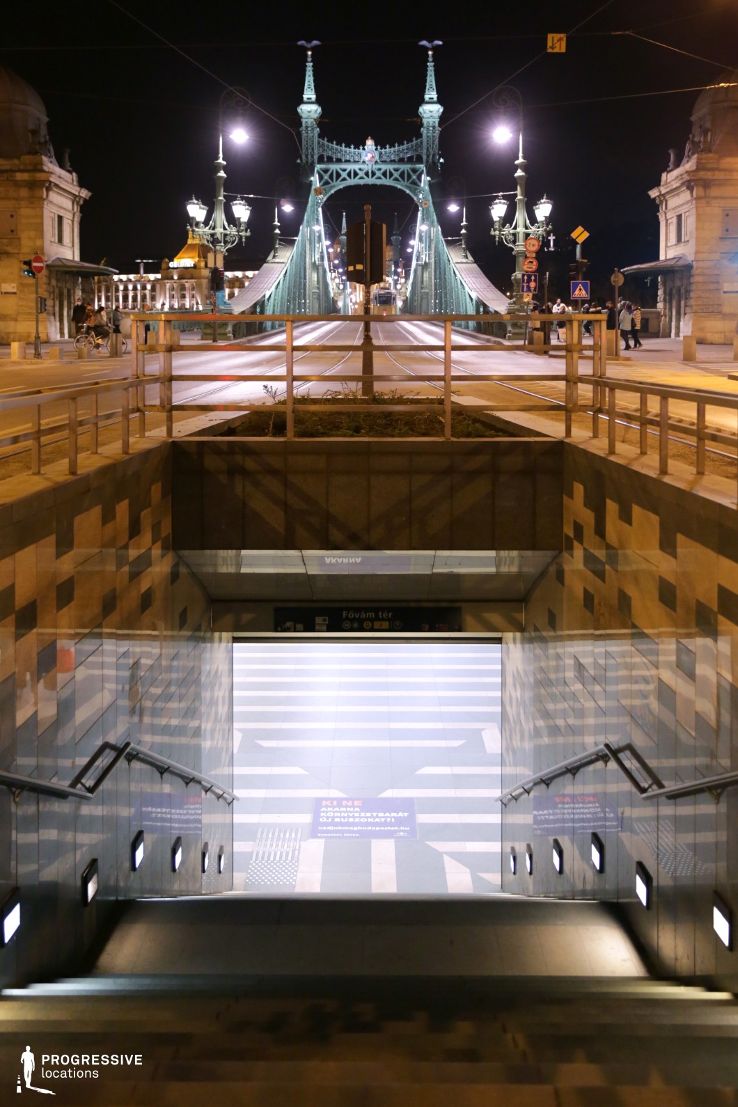 This striking urban scene captures the contrast between a modern underground metro entrance and a historic steel bridge, illuminated against the night sky. The symmetrical composition leads from the contemporary tiled stairway, with its sleek railings and geometric lighting, upward to the grand structure of the bridge, adorned with intricate ironwork and classical street lamps. The tram tracks and overhead cables add depth to the perspective, emphasizing the seamless integration of historic and modern transportation infrastructure. The warm glow of city lights enhances the architectural details, making this location ideal for capturing the dynamic energy of nighttime urban life.