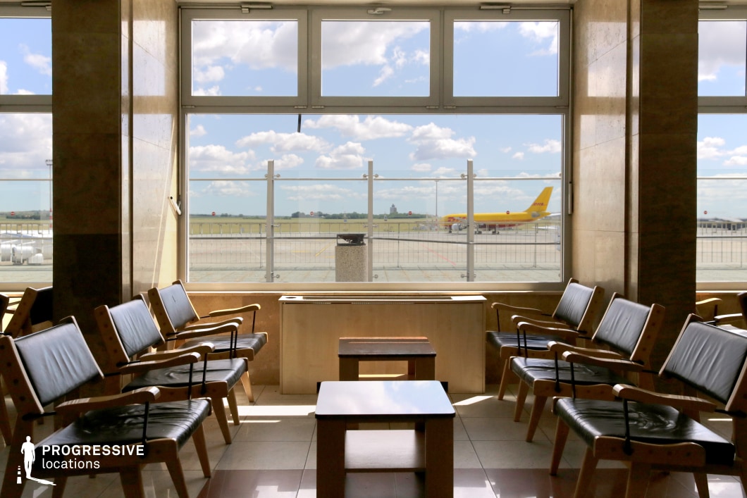 This waiting area inside an airport terminal features a mid-century modern aesthetic, characterized by its simple wooden furniture with black leather seating. Large floor-to-ceiling windows dominate the space, offering an expansive view of the tarmac where a yellow cargo plane is stationed in the background. The natural light streaming through the windows enhances the neutral tones of the polished stone flooring and walls, creating a warm yet functional atmosphere. The symmetrical arrangement of seating adds to the structured, orderly feel of the lounge, making it an ideal setting for scenes capturing air travel nostalgia, transit moments, or the quiet anticipation of journeys.