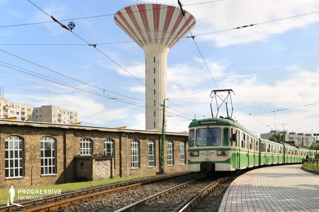 This image captures a green and white retro-style suburban train approaching a curved platform under a network of overhead cables. In the background, a distinctive water tower with red and white stripes dominates the skyline, adding an industrial yet iconic element to the scene. The adjacent brick depot, adorned with large arched windows and traces of graffiti, enhances the urban, slightly worn aesthetic. Surrounding residential buildings contribute to the metropolitan atmosphere, making this setting ideal for projects seeking an old-school railway ambiance, industrial cityscapes, or suburban transit visuals.