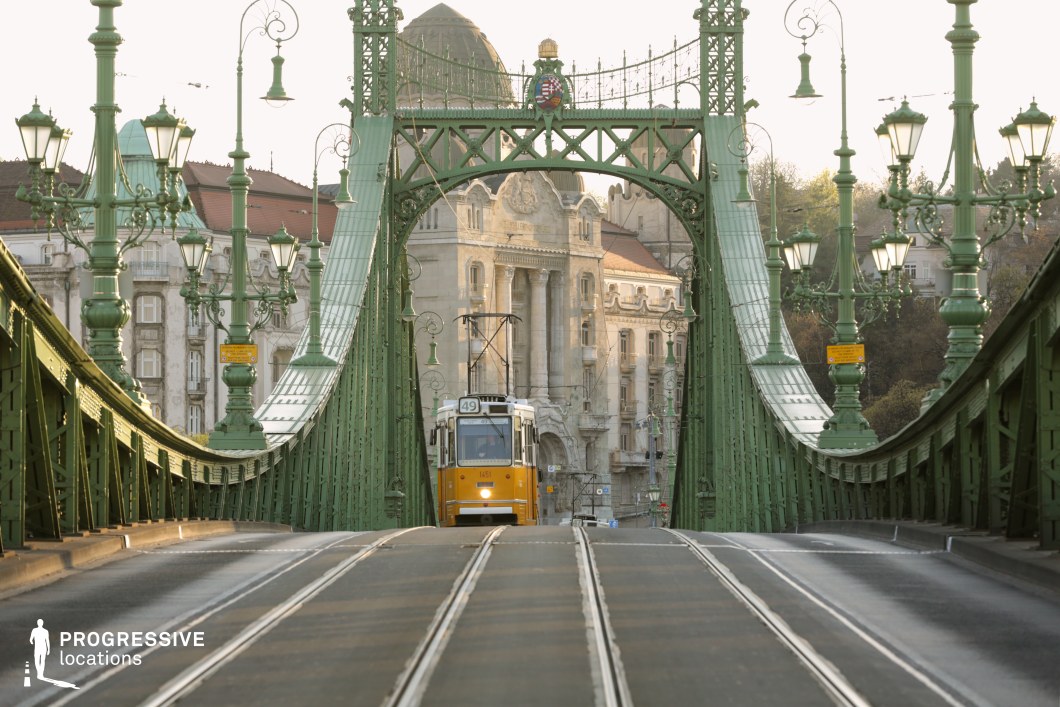 This striking image captures an ornate green wrought-iron bridge with a vintage yellow tram approaching in the distance. The bridge's intricate lampposts and decorative elements create a visually rich, historic atmosphere, while the tram’s presence adds a sense of urban movement and nostalgia. In the background, elegant historic buildings with domed rooftops and classical facades enhance the architectural grandeur of the scene. The slightly elevated tram tracks give a dynamic perspective, making this location ideal for cinematic cityscapes, period pieces, or projects highlighting European urban charm.