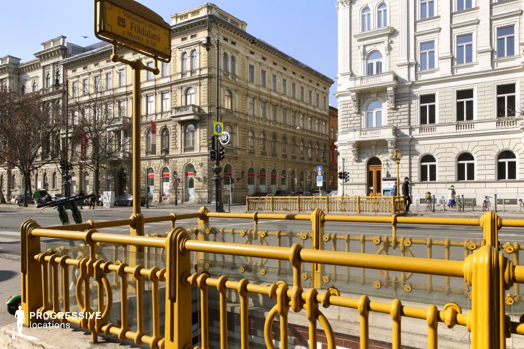 This image captures a beautifully preserved metro entrance with distinctive yellow wrought-iron railings, a signature element of early underground transport infrastructure. The metalwork frames a descending stairway, leading to an iconic metro system beneath a cityscape of elegant 19th-century buildings. The surrounding architecture showcases a mix of classical and neoclassical facades with arched windows and intricate stonework, evoking a grand European aesthetic. The balanced composition makes this location ideal for film projects, period pieces, or visuals emphasizing historic urban transport and architectural heritage.
