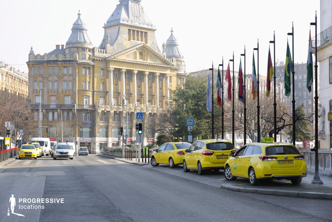 This image showcases a city intersection lined with yellow taxis in front of an imposing historic building featuring neoclassical and baroque influences. The structure’s ornate facade, grand columns, and intricate rooftop elements contribute to its monumental presence. A row of international flags adds a cosmopolitan atmosphere, while the urban setting includes modern traffic elements like pedestrian crossings and streetlights. The contrast between the historic architecture and contemporary transportation makes this location ideal for film scenes requiring a blend of old-world grandeur and modern city life.