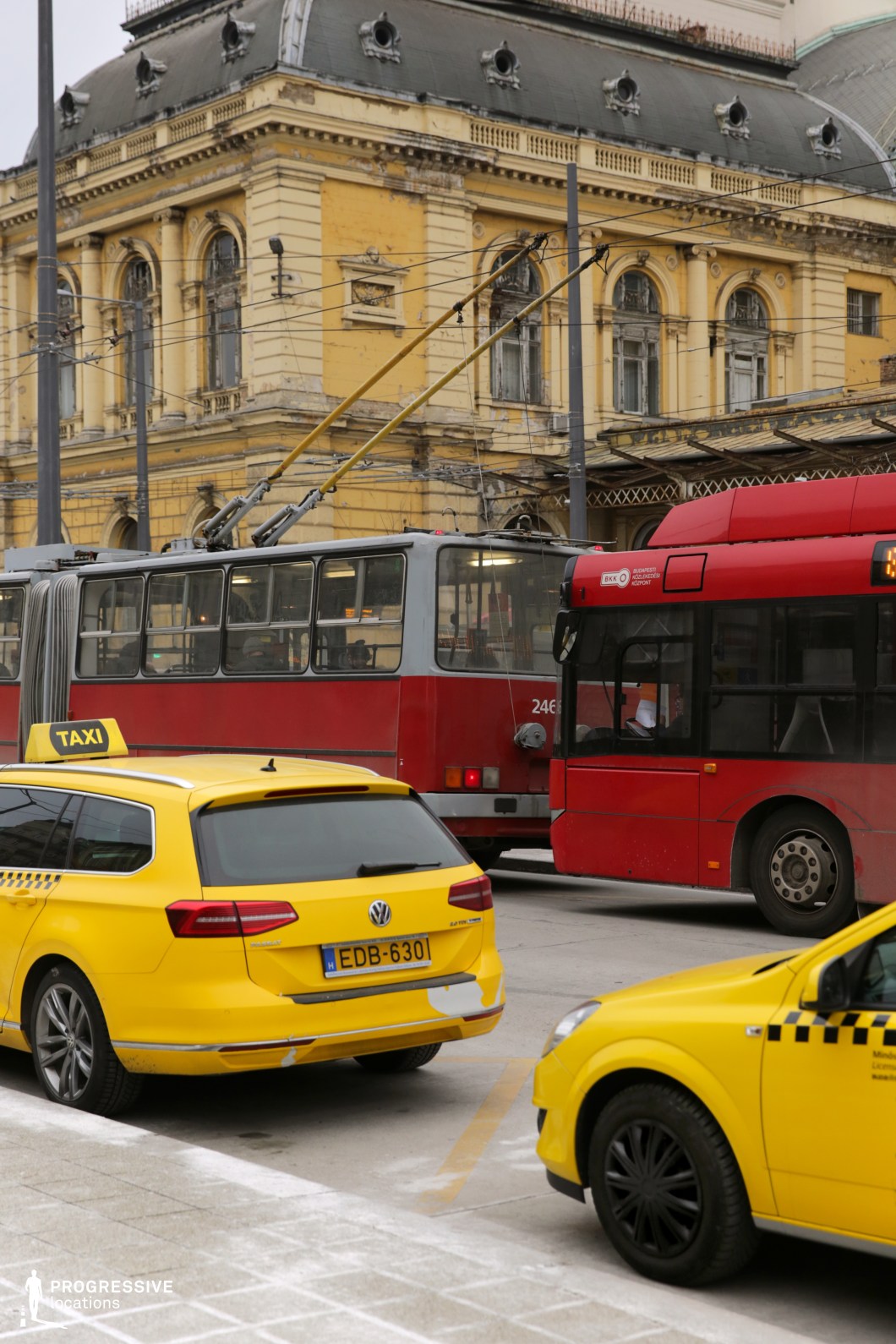 This image captures a dynamic city transport hub where modern and vintage elements intersect. A historic yellow building with intricate architectural details and a weathered facade serves as the backdrop for a bustling street filled with various modes of public transportation. A bright yellow taxi, a red trolleybus with overhead cables, and a red articulated bus occupy the foreground, creating a striking contrast of colors and vehicle types. The location suggests a well-connected urban setting, ideal for film scenes depicting city life, transportation networks, or historic European streetscapes.