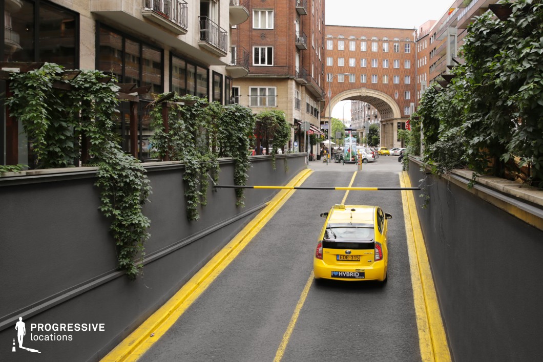 This image showcases a visually compelling cityscape featuring a yellow taxi moving through a narrow underpass lined with dark grey walls adorned with lush green ivy. The contrasting architectural styles are evident, with modern glass-fronted buildings on one side and traditional brick structures on the other. The focal point is the grand archway in the background, framing the lively street scene with cafes, pedestrians, and more yellow taxis in the distance. This location is ideal for urban storytelling, film scenes with a European city ambiance, or commercial projects emphasizing movement and city life.