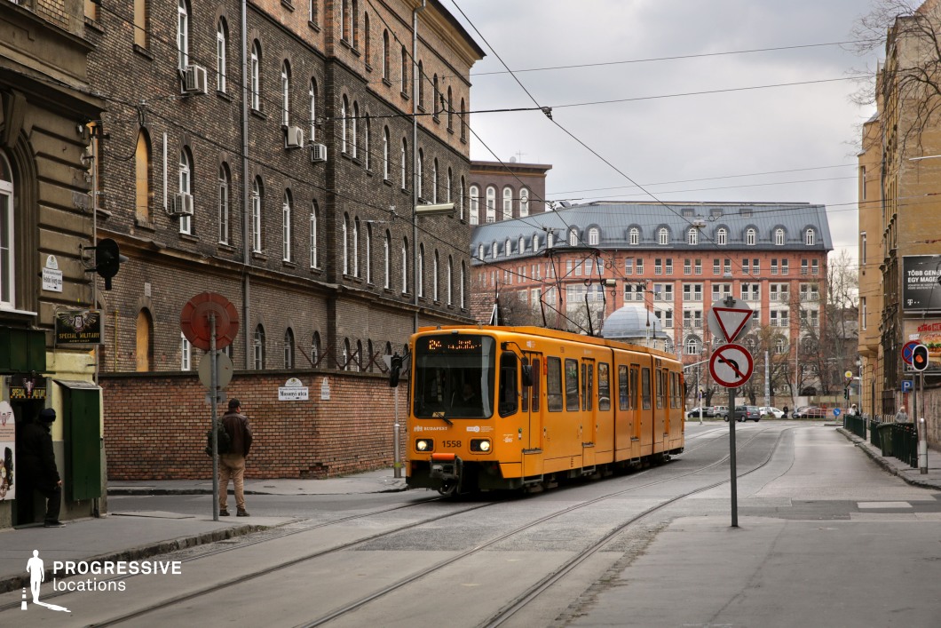 This image captures a striking urban scene where a yellow tram moves through a district dominated by historic brick architecture. The buildings feature arched windows, wrought-iron details, and signs of age that add character to the environment. Overhead tram cables crisscross the sky, enhancing the industrial aesthetic. In the background, a larger, more modern brick building with a mansard roof contrasts with the older facades. The muted tones of the overcast sky create a moody, atmospheric setting, making this location ideal for urban storytelling, period film settings, or projects requiring a European cityscape with depth and history.