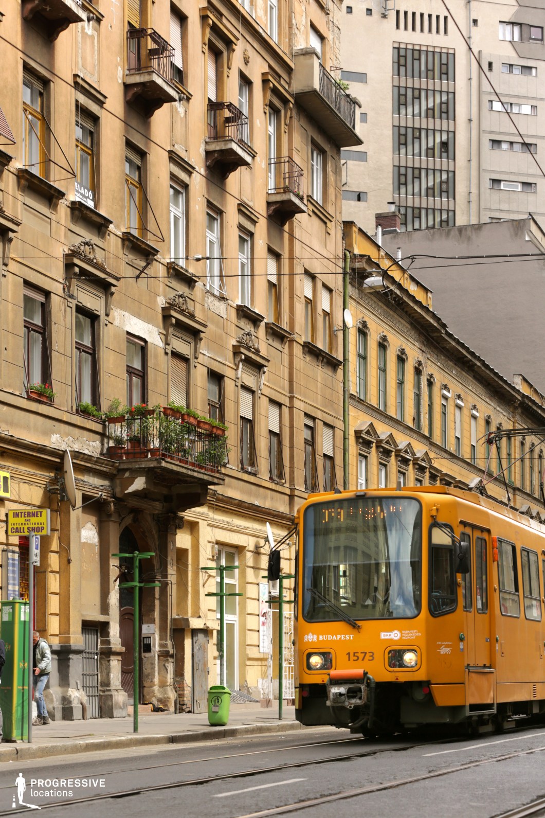 A vibrant city street lined with aged, classical-style buildings featuring ornate facades, decorative moldings, and small wrought-iron balconies. The warm, earthy tones of the structures, combined with peeling paint and weathered textures, emphasize a historic and slightly neglected aesthetic. The street is intersected by tram tracks, with an iconic yellow tram in motion, reinforcing the urban character and active public transportation system. In the background, a stark contrast emerges with modern, minimalist buildings, adding depth and visual tension between old and new architectural styles. The narrow street, overhead tram cables, and scattered storefronts create an intimate yet dynamic atmosphere, making this location ideal for film scenes set in a European metropolis, particularly for narratives that require an authentic, lived-in urban backdrop.