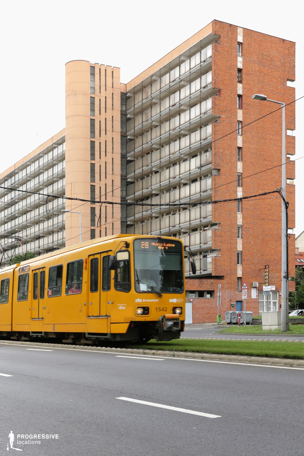 A striking urban environment dominated by a long, imposing socialist-era apartment building with a repetitive balcony design and a mix of brick and concrete textures. The architecture is defined by its functional, utilitarian aesthetic, featuring linear balconies and a curved stairwell tower that adds a subtle geometric variation. The foreground showcases a yellow tram in motion, emphasizing the city's well-integrated public transportation system. Overhead tram cables and streetlights contribute to the structured, metropolitan atmosphere. The contrast between the clean roadway and the weathered facade of the residential block creates a visually compelling juxtaposition, making this setting ideal for capturing a raw, post-socialist urban mood or a gritty, realist cityscape.