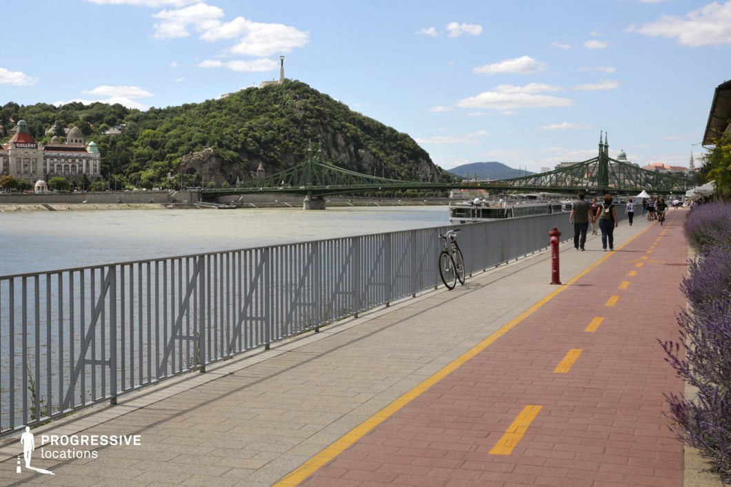 This image captures a well-maintained riverside promenade featuring a dedicated red-brick cycling and pedestrian path, bordered by a lavender-lined walkway. A sturdy metal railing separates the path from the water, leading the eye toward a striking green metal bridge in the background. Beyond the bridge, a lush hill with a monument at its peak rises dramatically, contrasting with the elegant historic buildings at its base. The scene balances urban and natural elements, creating a serene yet active city atmosphere. This location is ideal for film sequences that require a vibrant and picturesque European riverside setting, perfect for leisure scenes, travel montages, or contemplative moments against a scenic backdrop.