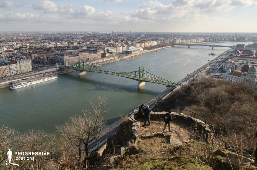 This aerial cityscape offers a panoramic view of an urban landscape intersected by a river, with a striking green metal bridge as a focal point. The vantage point from a rugged stone lookout showcases both the natural and architectural elements, blending historic and modern buildings along the waterfront. The river reflects the structures around it, while boats add movement to the composition. The foreground includes a rocky outcrop and sparse vegetation, enhancing the depth and framing the expansive view. Ideal for cinematic establishing shots or promotional visuals highlighting urban beauty and connectivity.