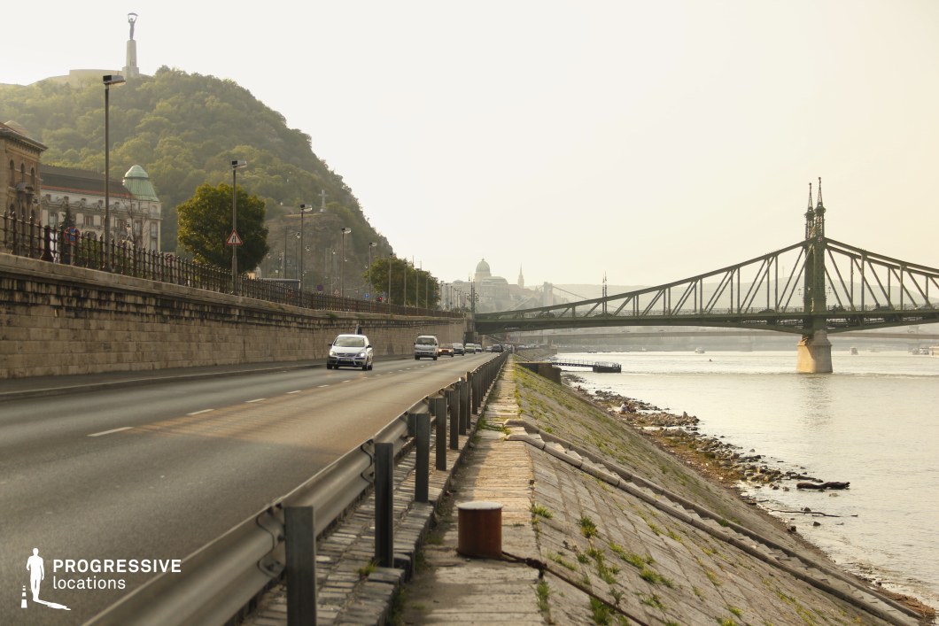 This image presents a sweeping riverside scene featuring a long, curving road running parallel to the water, bordered by a fortified stone wall with iron railings. The perspective leads towards a striking metal bridge with ornate spires, which elegantly spans the river, connecting both sides of the city. A towering green hill in the background adds natural contrast, crowned by a prominent monument. The embankment slopes gently down to the water, with patches of vegetation and scattered stones contributing to the raw, urban riverside aesthetic. The combination of historical and industrial elements makes this location ideal for dynamic film sequences, including chase scenes, reflective urban moments, or atmospheric establishing shots.