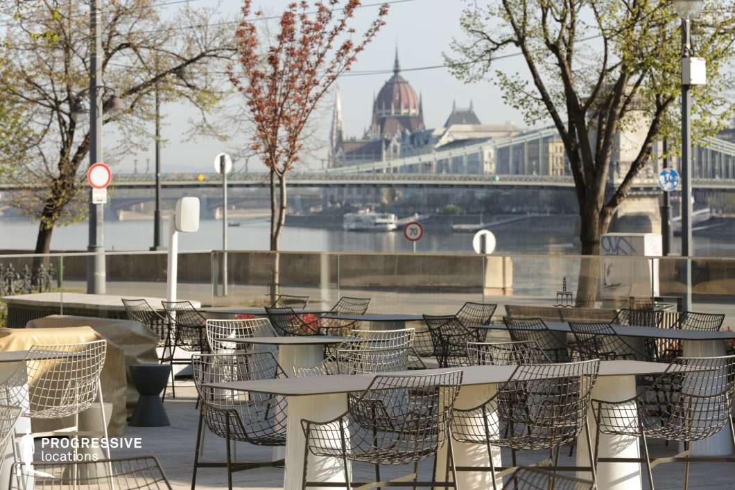 This image captures a stylish riverside café terrace with a breathtaking panoramic view of an iconic European cityscape. The foreground showcases a modern outdoor seating area with contemporary wireframe chairs and sleek tables, set against a glass barrier that maintains an open and unobstructed perspective. The background features a striking historic domed building and an elegant suspension bridge spanning the river, blending architectural grandeur with urban vitality. Trees with budding leaves frame the scene, adding a natural touch to the refined setting. This location is ideal for film sequences requiring a sophisticated yet relaxed atmosphere, perfect for conversations, introspective moments, or elegant travel montages with a European charm.
