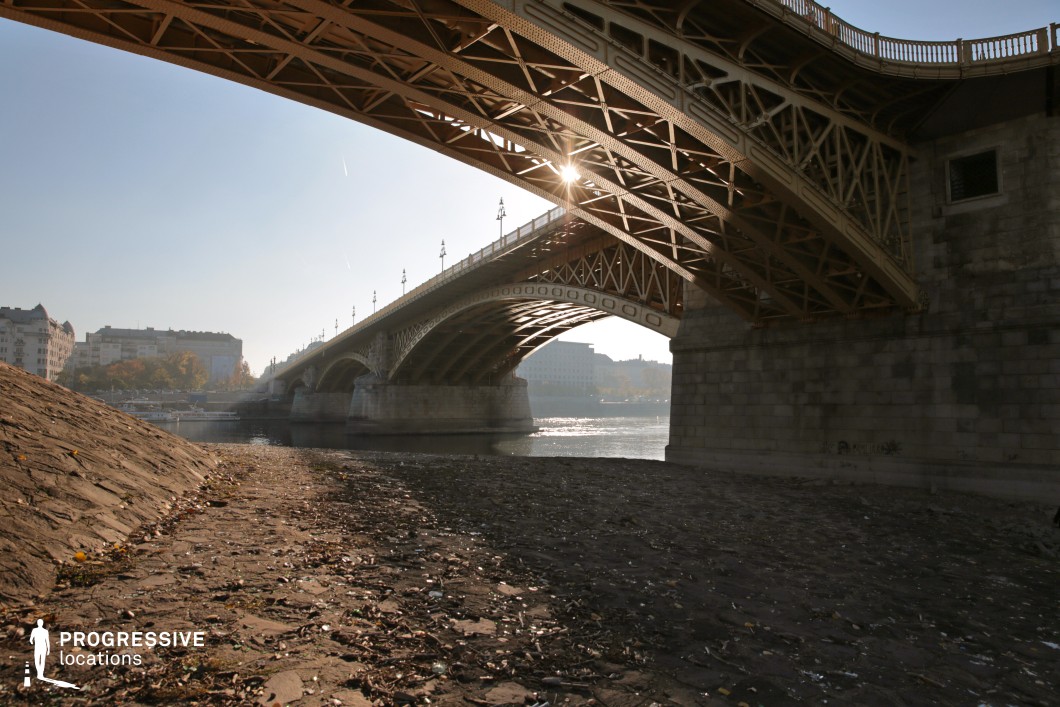 This image presents a dramatic perspective from beneath a grand historic bridge, emphasizing the intricate cast-iron framework and sturdy stone pillars. The sun peeks through the metal structure, casting dynamic shadows and adding depth to the composition. The riverbank in the foreground, textured with scattered debris and weathered stone, contrasts with the structured elegance of the bridge. In the background, classic city buildings line the river, reinforcing the blend of history and urban development. This setting is ideal for film scenes that require a strong architectural presence, evoking a sense of mystery, nostalgia, or urban grandeur—perfect for dramatic encounters, contemplative moments, or visually striking establishing shots.