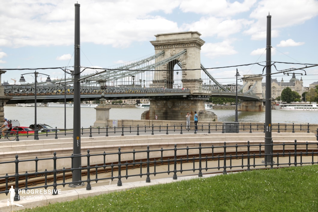 The image presents a view of the iconic Chain Bridge, with its grand suspension structure spanning across the Danube River. The foreground features neatly aligned tram tracks that curve along the riverbank, leading toward the bridge, while elegant lampposts and railings line the path, adding a classic touch to the scene. The natural green space and the well-maintained urban setting highlight the balance between the historic architecture of the bridge and the modern infrastructure. This location offers a blend of iconic architecture and functional urban elements, making it ideal for capturing both historic and contemporary cityscapes.