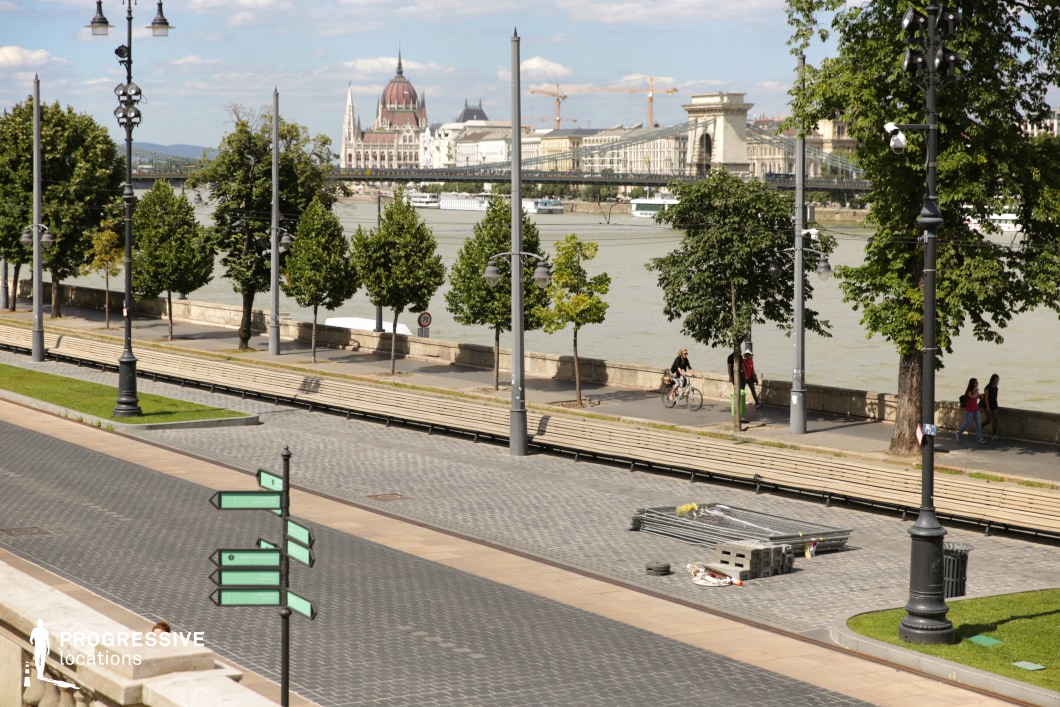 This image captures an elegant riverside promenade framed by lush trees, vintage-style lampposts, and well-maintained cobblestone pavement. The open and structured design of the walkway includes long wooden benches and a decorative green directional sign, adding a sense of navigation and movement. In the background, a grand European cityscape is visible, featuring an iconic domed building, ornate spires, and a historic suspension bridge that enhances the depth of the composition. The calm river reflects the architectural beauty, emphasizing the balance between urban sophistication and natural elements. This setting is ideal for film scenes requiring a refined European backdrop, suitable for contemplative moments, leisurely city strolls, or establishing shots in historical or travel-based narratives.