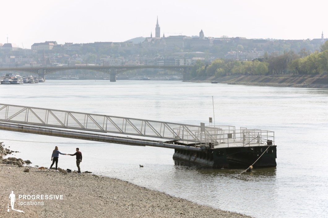 This image captures a quiet riverside setting with a floating dock extending into calm waters, framed by a gently sloping rocky shoreline. In the background, a historic cityscape rises over the opposite bank, featuring a mix of architectural styles, including spires and fortified structures. A bridge spans the river, connecting the urban areas on either side. The dock’s metallic structure and angular lines contrast with the natural elements of the water and greenery. This location is ideal for film scenes depicting moments of solitude, contemplation, or transitional storytelling elements, such as departures, arrivals, or reflective interludes.