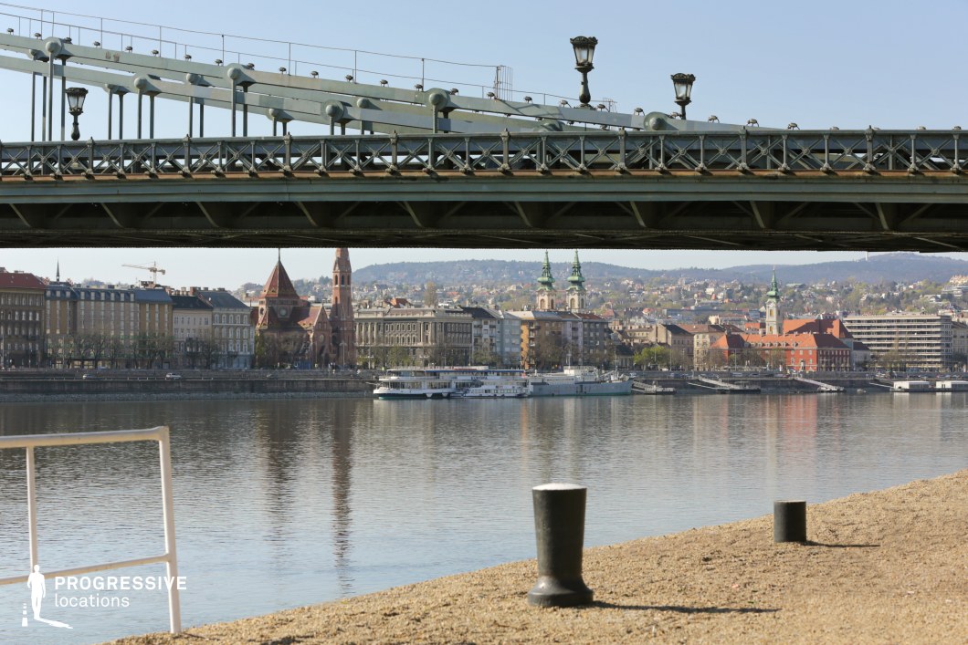 This image presents a striking perspective from beneath an iron bridge, framing a picturesque riverside cityscape. The bridge's intricate metal framework and vintage lampposts contrast with the calm waters below, creating a layered composition that adds depth and symmetry. In the background, a mix of historic and modern architecture lines the waterfront, including red-bricked churches with towering spires and baroque-style buildings with green rooftops, set against rolling hills. The foreground features a textured, sandy dock with metal mooring posts, reinforcing the setting’s connection to river transport. This location offers a versatile backdrop for film scenes that require a European riverfront ambiance, ideal for suspenseful urban dramas, contemplative character moments, or visually rich establishing shots.