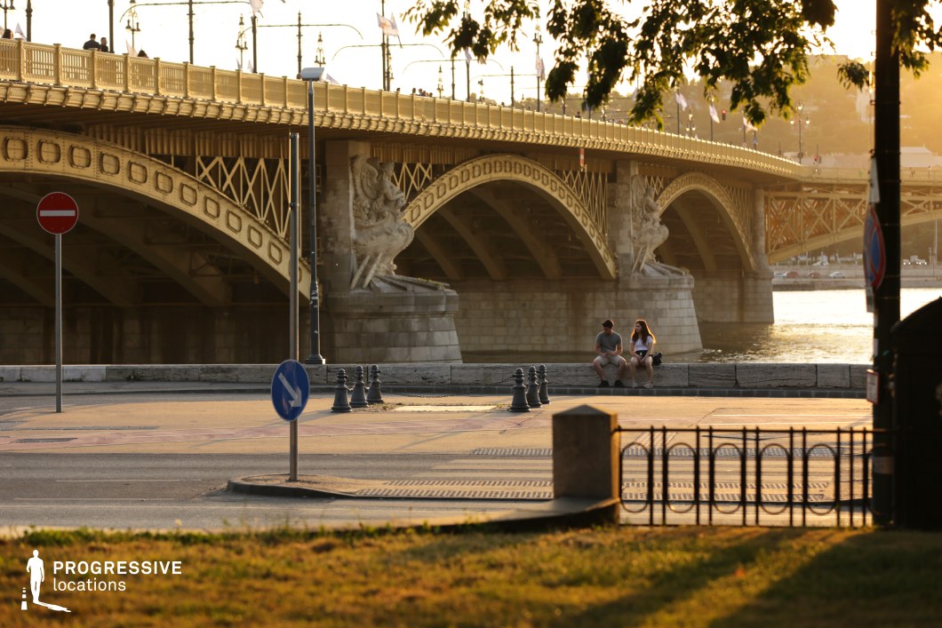 The image captures a striking view of a historic yellow bridge spanning the Danube River, highlighted by its intricate arches and decorative stone sculptures at its base. The warm sunlight casts a golden glow over the scene, emphasizing the bridge's architectural details. The foreground features a spacious urban area with roads and traffic signs, adding to the blend of historical and modern elements. The surrounding environment, with its clean lines and simple structures, complements the grandeur of the bridge, making this location ideal for capturing both the historic charm and contemporary atmosphere of the city.