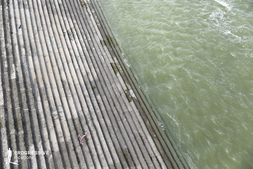 This image captures a striking riverside composition, featuring a vast set of stone steps descending directly into the flowing water. The repetitive linear patterns of the weathered stone create a strong geometric effect, emphasizing depth and perspective. Small patches of vegetation growing between the steps add texture and a sense of age to the structure. The contrast between the solid, immovable stone and the ever-moving water enhances the visual tension of the scene. This location is ideal for film sequences requiring a dramatic, contemplative, or symbolic setting, perfect for moments of solitude, introspection, or transitions between urban and natural environments.