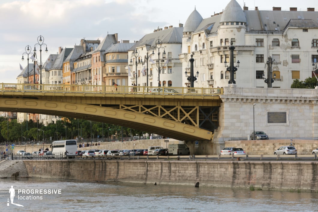 This image captures a harmonious blend of elegant architecture and infrastructure along a European riverside. The foreground features a striking yellow iron bridge with intricate detailing, supported by a sturdy stone base, seamlessly connecting both sides of the city. Ornate lampposts line the bridge, reinforcing its historic charm. Beyond the bridge, a row of architecturally diverse buildings, characterized by mansard roofs, turrets, and classic facades, adds visual depth and a sense of history. The riverbank below, with its stone embankment and roadway, introduces an element of urban activity. This setting is ideal for film scenes that require a rich, historic backdrop with a touch of urban vibrancy, perfect for establishing shots, European period pieces, or sophisticated city narratives.