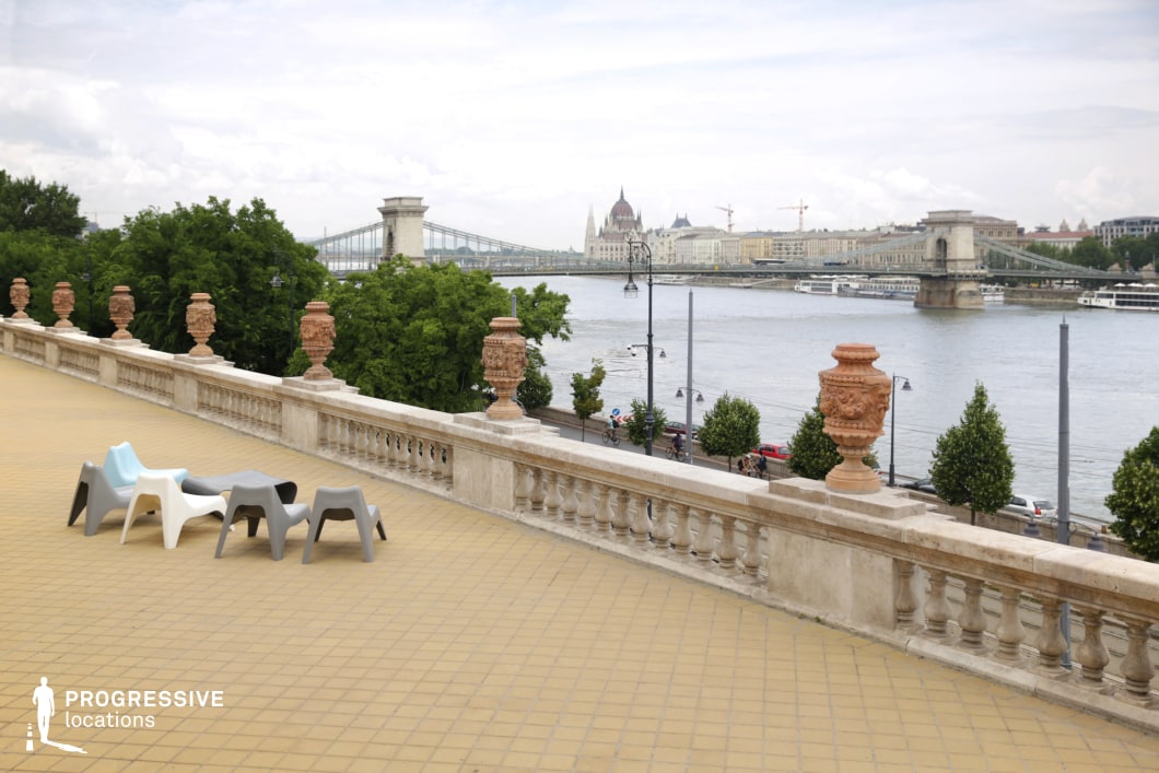 The image features a wide terrace adorned with a historic balustrade, offering a stunning view of the Chain Bridge and the Hungarian Parliament across the Danube River. The terrace is spacious, with modern chairs in contrasting colors, adding a contemporary touch to the classic surroundings. The lush greenery and neatly arranged trees along the riverbank further enhance the peaceful atmosphere. The combination of the elegant balustrade, the modern seating, and the scenic riverfront creates a harmonious blend of tradition and modernity, making this location ideal for leisurely outdoor settings or capturing expansive city views.