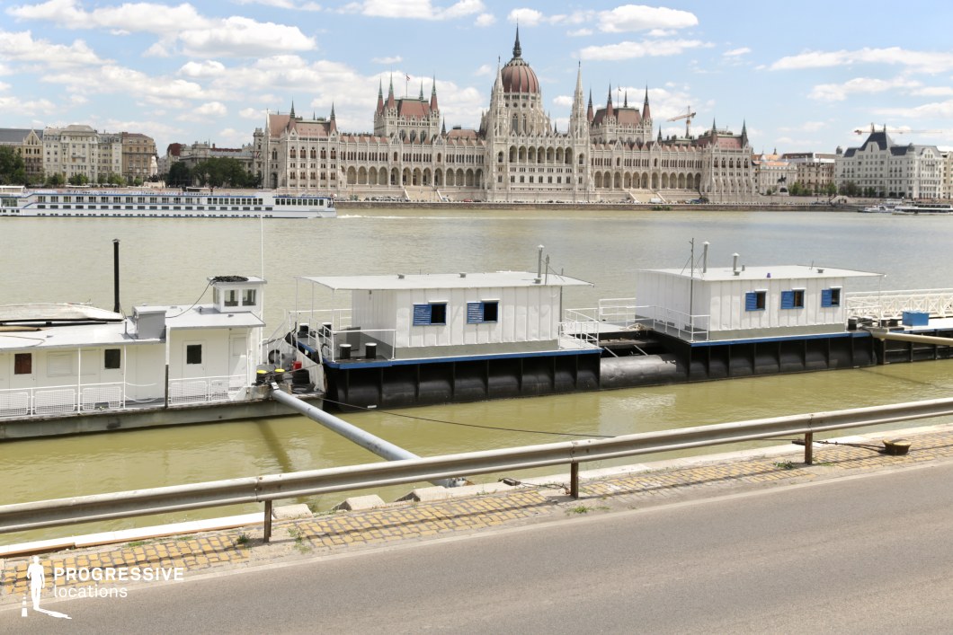 This image presents a striking contrast between industrial river infrastructure and grand historic architecture. In the foreground, a set of floating dock structures and moored boats with utilitarian white exteriors line the riverbank, anchored near a guardrail and a paved roadside. The river, central to the composition, reflects the light and creates a visual divide between the functional maritime elements and the ornate, neo-Gothic parliamentary building in the background. The symmetrical façade, adorned with spires and intricate details, dominates the skyline, reinforcing the grandeur of the cityscape. This location is ideal for film scenes that juxtapose urban industry with historical elegance, making it suitable for establishing shots, political dramas, or contemplative waterfront sequences.