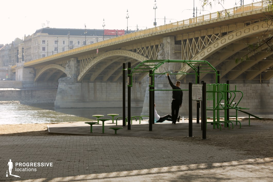 This image presents a unique blend of urban recreation and historic architecture, featuring a modern outdoor gym structure in the foreground with a grand yellow iron bridge arching over the river in the background. The green workout equipment, with its angular and functional design, contrasts with the ornate, classical detailing of the bridge’s lampposts and sculptural elements. The paved riverside setting offers an open and airy atmosphere, while the water and distant city buildings add depth to the composition. This location is ideal for film scenes that juxtapose modern urban life with historical surroundings, making it suitable for dynamic training montages, reflective city moments, or energetic outdoor sequences in a European setting.