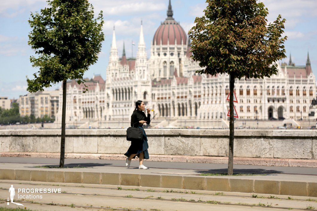 This image captures a striking contrast between a lush, tree-lined pedestrian walkway and an iconic, neo-Gothic architectural masterpiece in the background. The paved path is bordered by evenly spaced young trees, creating a natural frame for the monumental building behind. A weathered stone balustrade separates the foreground from the river, adding depth and a sense of urban history. The grand structure in the background, with its intricate spires and domed roof, dominates the skyline, exuding a regal and authoritative presence. The scene balances greenery and historic grandeur, making it an excellent setting for elegant, contemplative, or diplomatic film sequences, particularly those requiring a refined European city atmosphere.