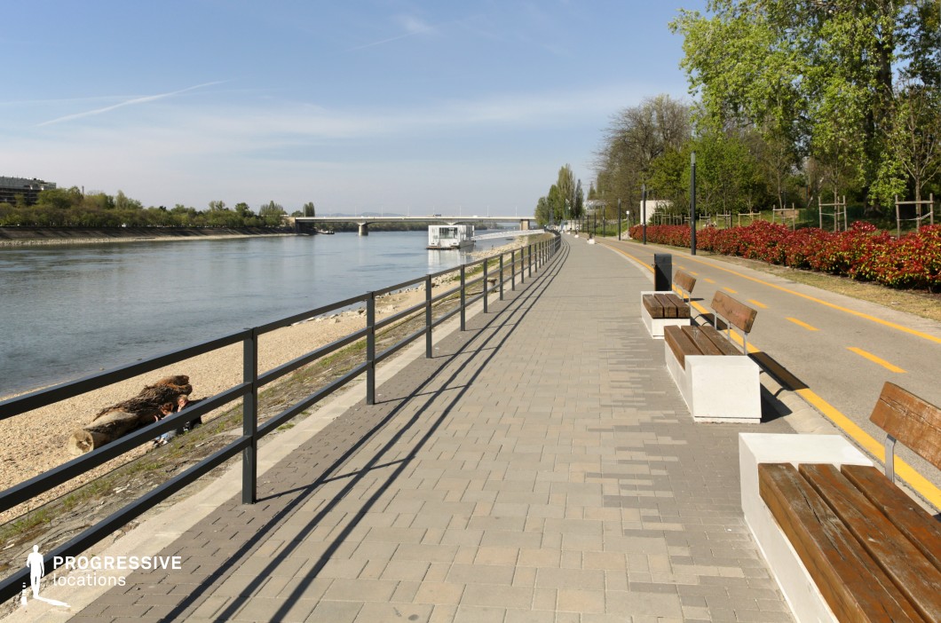 This image showcases a well-maintained riverside promenade with a paved walkway lined by modern wooden benches set on concrete bases. A parallel bicycle lane with bright yellow markings runs alongside, bordered by neatly arranged red-leafed shrubs and tall trees that provide a natural touch to the urban design. A black metal railing separates the path from the sandy shoreline, where logs and natural debris are scattered. In the distance, a sleek pedestrian bridge spans the river, enhancing the depth of the scene. This location offers a clean, contemporary aesthetic ideal for lifestyle photography, urban planning visuals, or scenic backdrop needs in film productions.
