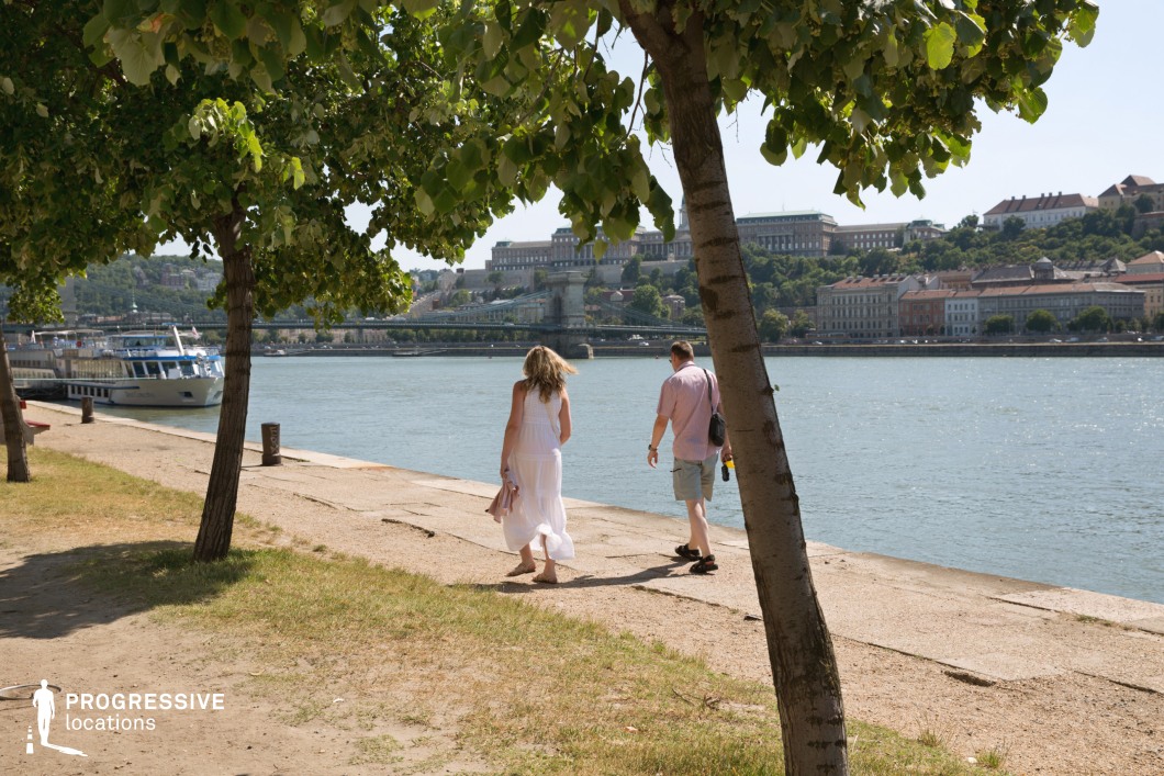 This image showcases a scenic riverside promenade shaded by lush green trees, with a calm waterway stretching towards a historic cityscape in the background. The composition is framed by tree trunks, adding depth and perspective to the open pathway leading along the riverbank. A grand suspension bridge connects both sides of the city, while elegant buildings and a domed palace sit atop a hill, reinforcing the area's architectural richness. The presence of moored boats adds a touch of movement and urban life to the serene atmosphere. This location is ideal for film sequences requiring a tranquil yet sophisticated European riverside setting, perfect for contemplative moments, leisurely strolls, or establishing shots that highlight historic and scenic beauty.