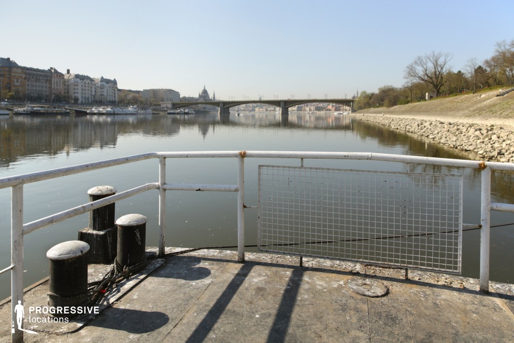 This image captures a riverside pier with weathered metal railings and mooring bollards in the foreground, offering an industrial contrast to the serene water beyond. The calm river reflects the surrounding architecture, including historic and modern buildings lining the waterfront. In the distance, a grand bridge spans the river, connecting both sides of the city, while a tree-lined embankment with a rocky shoreline adds a natural element to the composition. The scene provides a dynamic yet tranquil urban setting, ideal for cinematographic use or promotional visuals related to riverfront developments and city life.