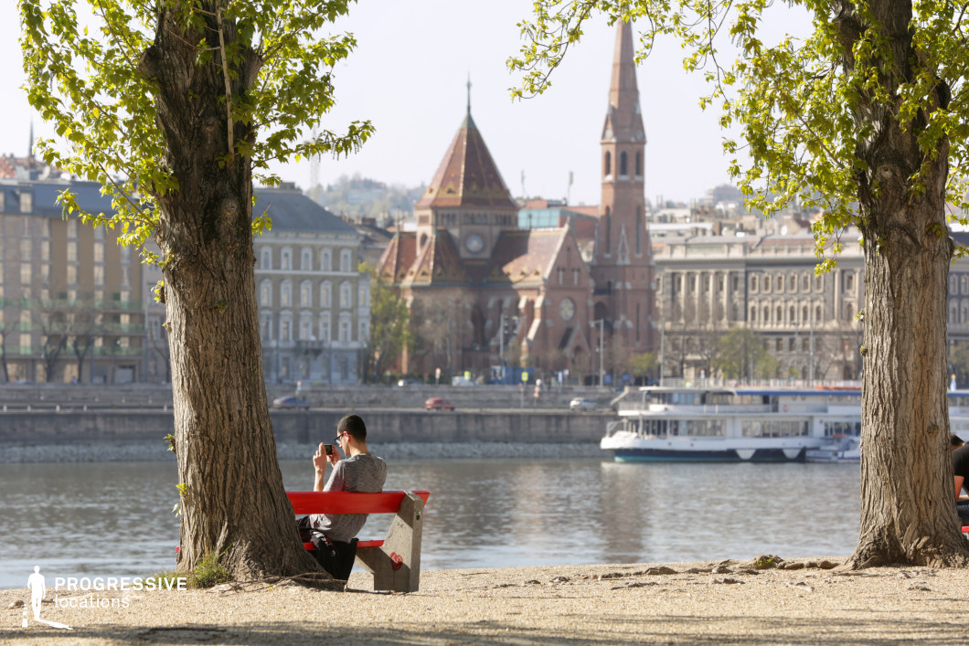 This image presents a serene riverside setting framed by two mature trees, drawing attention to a striking architectural backdrop. A vibrant red bench in the foreground adds a bold contrast to the earthy tones of the gravel and tree trunks. Across the river, an ornate red-brick church with pointed spires and intricate detailing stands prominently, surrounded by elegant classical buildings. The calm waters reflect the cityscape, while boats docked along the shore add a sense of movement and urban life. This location is ideal for film scenes requiring a peaceful yet visually rich European setting, perfect for contemplative moments, character-driven narratives, or atmospheric establishing shots.