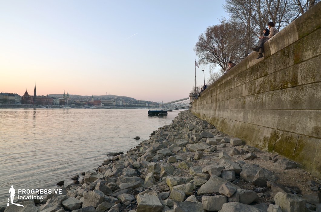 This image captures a rugged riverside embankment lined with large, uneven rocks, leading toward a sturdy, weathered stone wall. The elevated walkway above the embankment provides a striking contrast to the raw texture of the shoreline below. Across the river, a historic cityscape with pointed church spires and elegant buildings stretches along the waterfront, backed by rolling hills. The soft hues of the sky and the calm water create a tranquil yet cinematic atmosphere. This location is ideal for film scenes requiring a balance between urban and natural elements, perfect for introspective moments, atmospheric establishing shots, or sequences that emphasize solitude and contemplation.