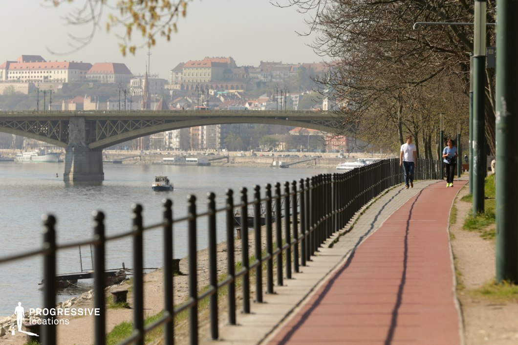 This image captures a scenic riverside pathway lined with black wrought iron railings, offering a structured yet inviting environment for pedestrians and joggers. The red-paved running path follows the curve of the embankment, bordered by trees that provide a natural canopy. In the background, a grand bridge spans the river, connecting both sides of the city, while boats glide along the water, adding to the dynamic urban setting. The historic architecture on the opposite bank enhances the location's visual appeal, making it a versatile backdrop for film productions, lifestyle photography, or travel-focused projects.